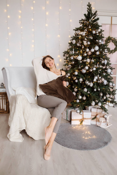 Cheerful woman sitting on the chair next to Christmas tree, celebrating winter holidays at home.  - Foto, imagen