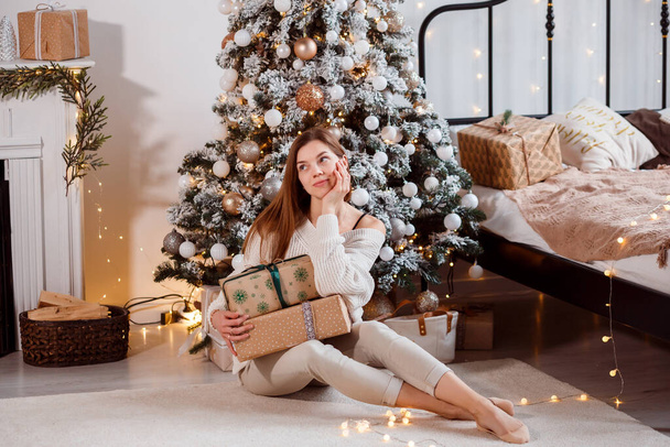 Romantic woman with gift boxes sitting next to Christmas tree. Celebrating winter holidays at home - 写真・画像