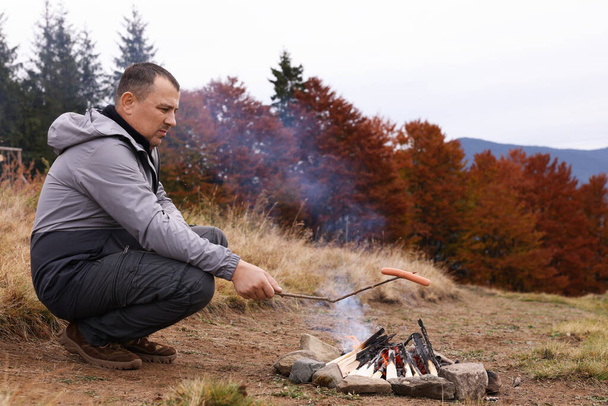 Man roasting tasty sausage over campfire near forest. Camping season - Fotoğraf, Görsel