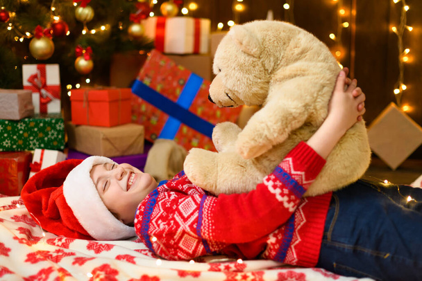 Kind meisje poseren in nieuwjaar of kerst decoratie. Feestelijke lichtjes en veel cadeaus, een elegante kerstboom met speelgoed. Het meisje draagt een rode trui en een kerstmuts. Ze speelt met een beer. - Foto, afbeelding