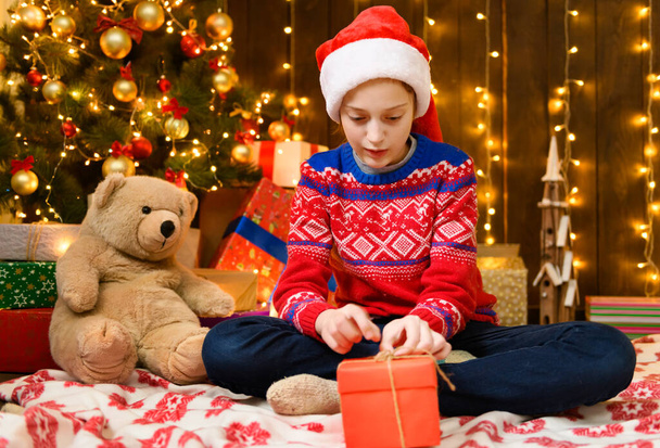 Child girl posing in new year or christmas decoration. Holiday lights and lots of gifts, an elegant Christmas tree with toys. The girl is wearing a red sweater and a Santa hat, she opening a gift box - Foto, imagen