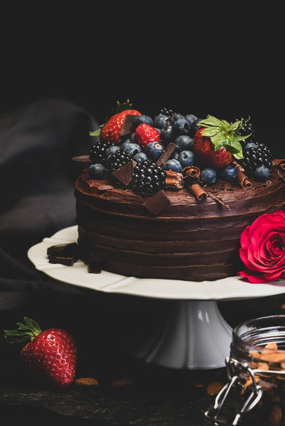 Delicious Double Chocolate Cake Dessert topped with Chocolate Flakes and Summer Fruits, including blackberries, strawberries and blueberries. Styled on a white cake stand with a rose and jar of almonds against a dark background on stone - Zdjęcie, obraz
