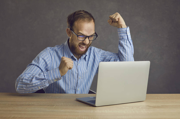 Excited businessman sitting at table with laptop computer, fist pumping and shouting Yes - Фото, изображение