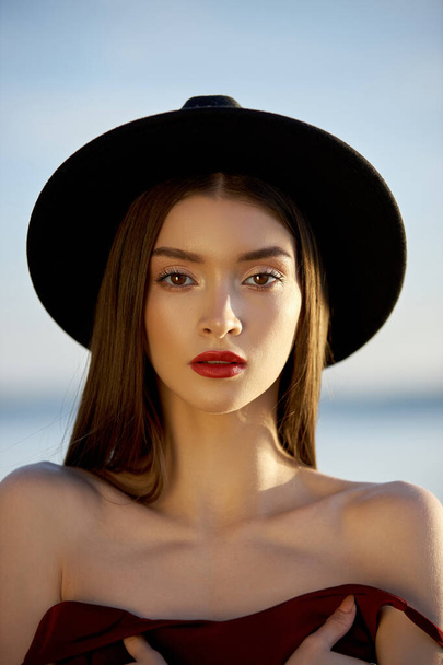 Portrait of a beautiful woman with long hair in a big round black hat. Beauty skin face, bare shoulders. Girl golden light at sunset - Foto, Imagem