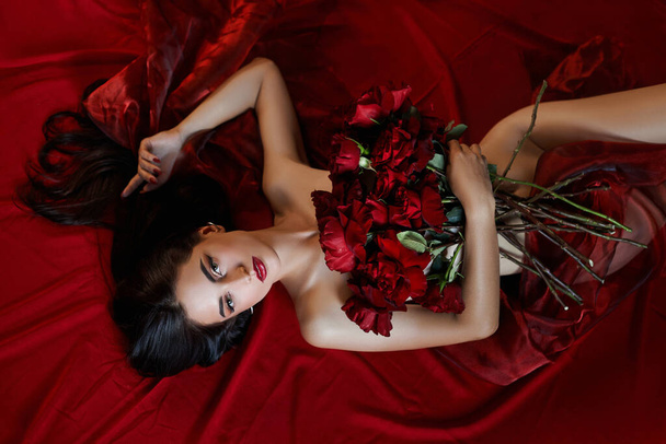 Beautiful sexy brunette with a bouquet of red roses lying on the floor, naked body parts, erotic portrait of a woman - Фото, изображение