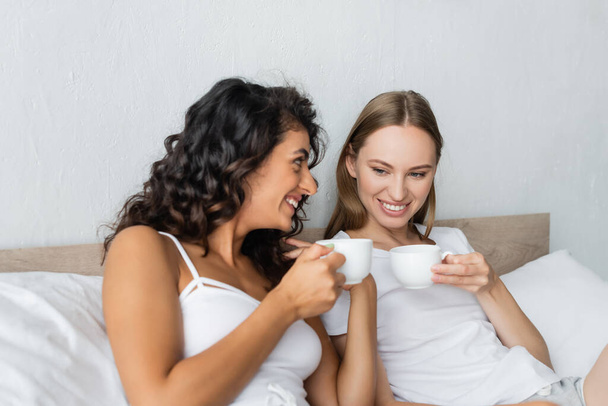 cheerful lesbian couple holding cups of coffee in bed - Фото, зображення