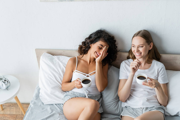 high angle view of joyful lesbian couple holding cups of coffee in bed - Fotó, kép