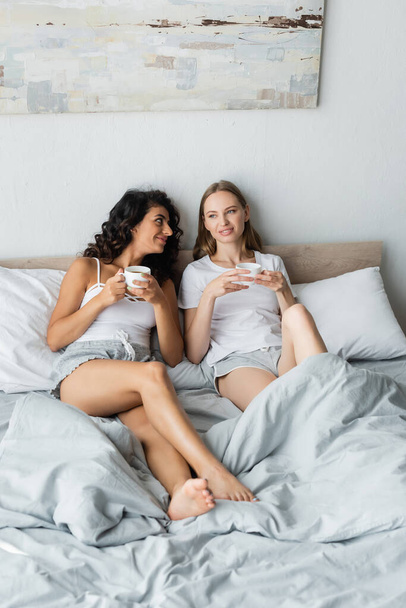 pleased young lesbian couple holding cups of coffee in bed - Foto, Bild