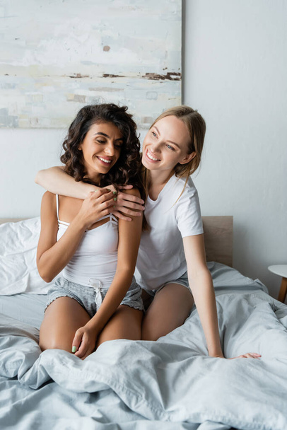 joyful lesbian women hugging in bedroom  - Фото, изображение
