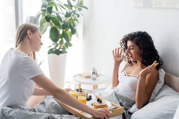 young woman bringing tray with breakfast to excited girlfriend in bed - Foto, immagini