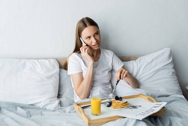 jovem mulher falando no smartphone perto da bandeja com café da manhã no quarto  - Foto, Imagem
