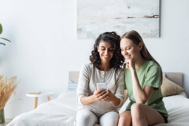 pregnant woman using smartphone near happy girlfriend in bedroom - Photo, Image