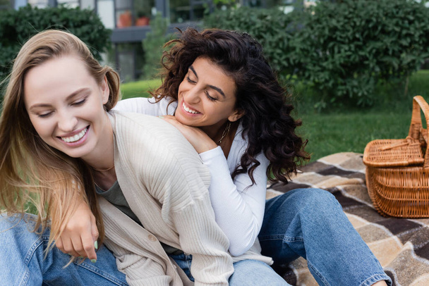 curly lesbian woman hugging happy girlfriend on checkered blanket - Φωτογραφία, εικόνα