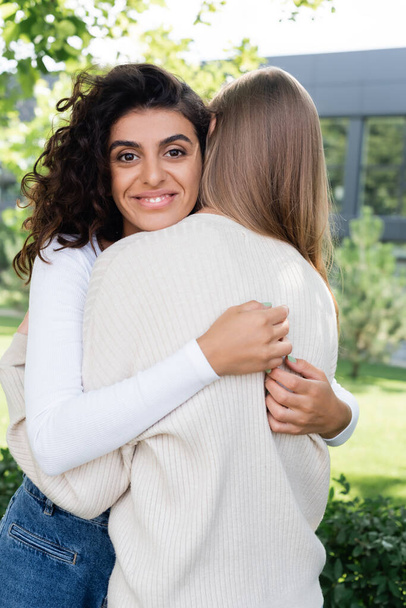 Χαρούμενη σγουρή γυναίκα αγκαλιάζει φίλη έξω - Φωτογραφία, εικόνα