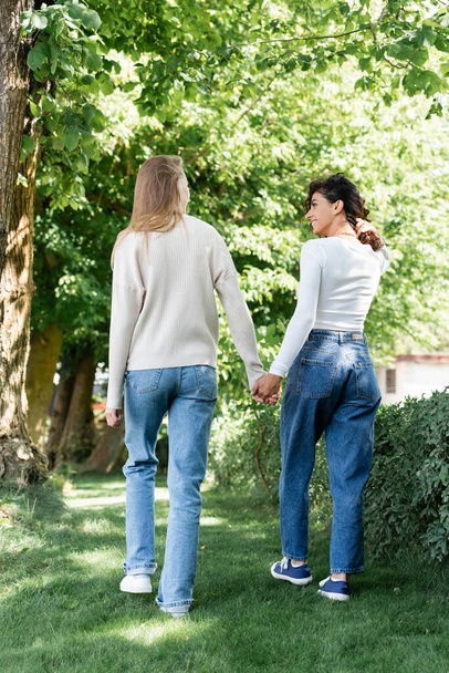 full length of pleased lesbian couple in jeans holding hands and walking outside  - Foto, imagen