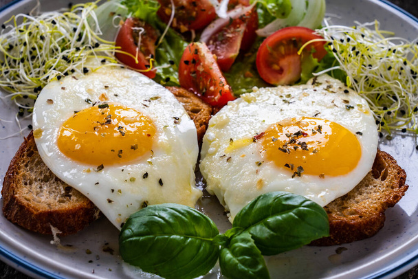 Sunny side up eggs on toasted bread, parmesan and fresh vegetables on wooden table  - Photo, image