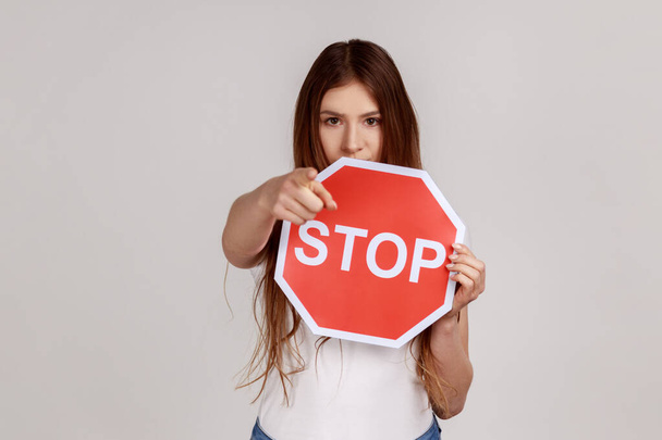 Serious woman holding big red stop road sign and pointing finger on you looking at camera with strict expression, warning, wearing white T-shirt. Indoor studio shot isolated on gray background. - Фото, зображення