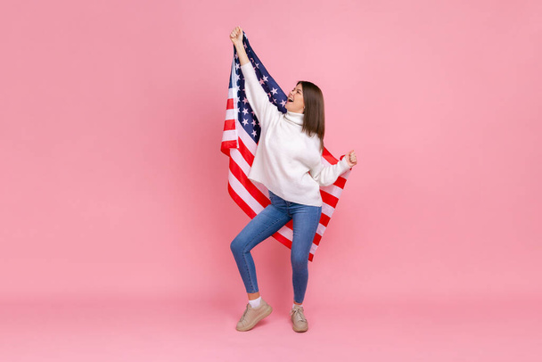Full length photo of woman raised arms, holding american flag, celebrating national holiday, dancing, wearing white casual style sweater. Indoor studio shot isolated on pink background. - Photo, image