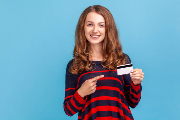 Smiling woman wearing striped casual style sweater, points finger at credit card, looking with toothy smile, recommend bank or shopping discounts. Indoor studio shot isolated on blue background. - Foto, Imagem