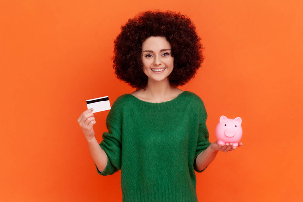 Positive kind young adult woman with Afro hairstyle wearing green casual style sweater standing showing to camera piggy bank and credit card. Indoor studio shot isolated on orange background. - Fotó, kép