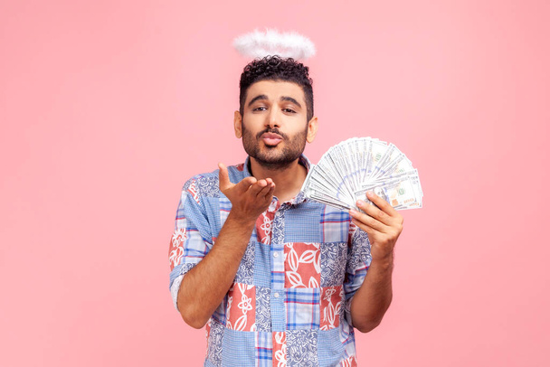 Portrait of romantic bearded man wearing blue casual style shirt with nimbus on head sending air kissing to camera, holding dollars bills. Indoor studio shot isolated on pink background. - Fotoğraf, Görsel