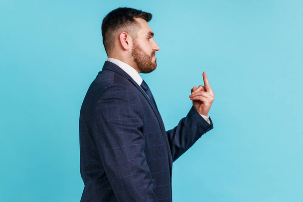 Profile portrait of bearded aggressive hater, angry man wearing official dark style suit showing middle finger, rude gesture of disrespect. Indoor studio shot isolated on blue background. - Foto, afbeelding