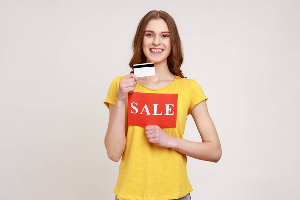 Portrait of young female with brown hair wearing yellow T-shirt showing credit card and sale sign, shopping cash back, looking at camera with smile. Indoor studio shot isolated on gray background. - Foto, imagen