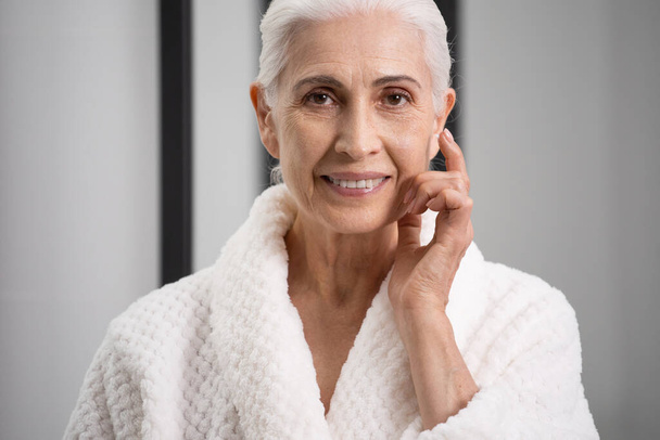 Portrait of smiling aged woman in robe. Anti-aging concept. Self-care at home. Idea of loving yourself. Female positive pensioner in bathroom - Photo, Image