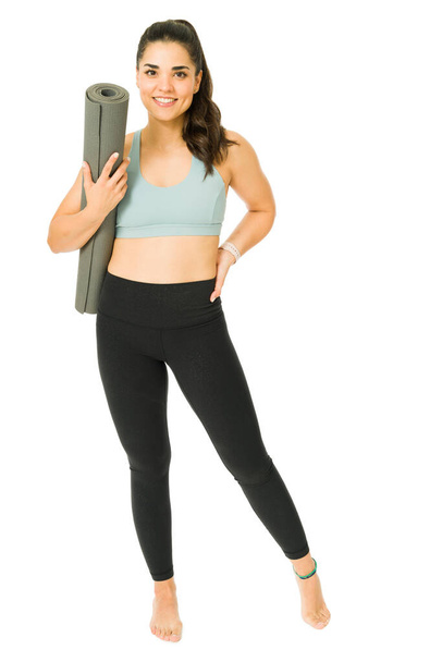 Valmiina treeneihini. Urheilullinen iloinen nainen, jolla on joogahousut hymyillen ja kantaen harjoitusmattoa  - Valokuva, kuva