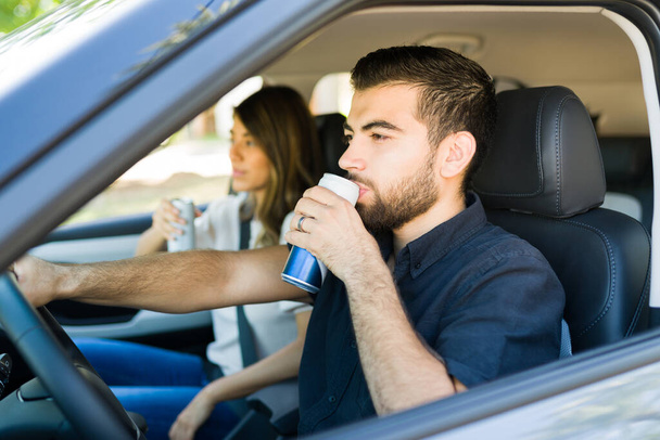 Thirsty young man drinking a beverage and driving a car with his girlfriend while traveling together - Zdjęcie, obraz