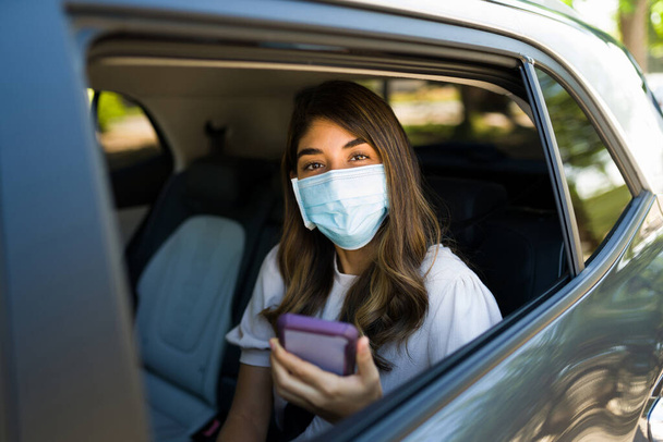 Hispánská mladá žena s obličejovou maskou při pohledu do kamery při jízdě sdílet auto během pandemie - Fotografie, Obrázek