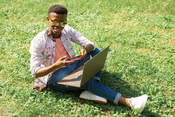 studente africano seduto sull'erba con computer portatile e telefono - Foto, immagini