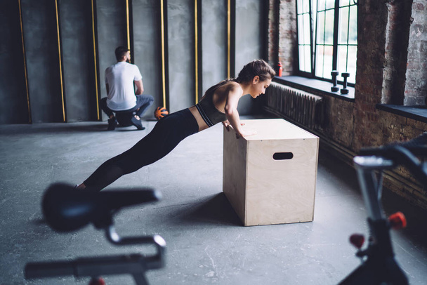 Side view of full length of focused sportswoman in activewear doing push ups from box during workout in spacious gym with fitness trainer - Foto, imagen