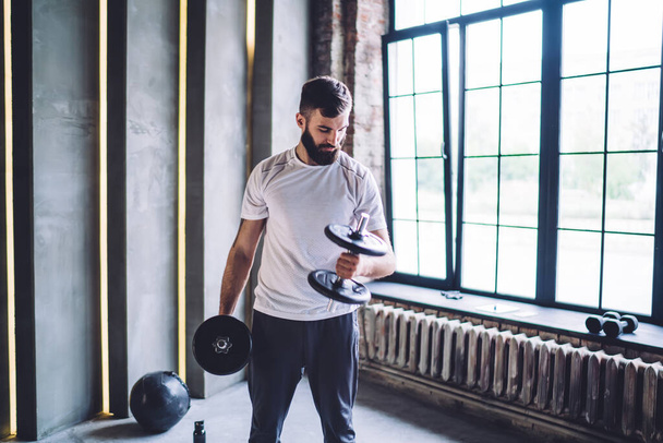 Young bearded male in sportswear using dumbbells for strength training to develop muscles while workout in daytime in modern gym - Foto, imagen