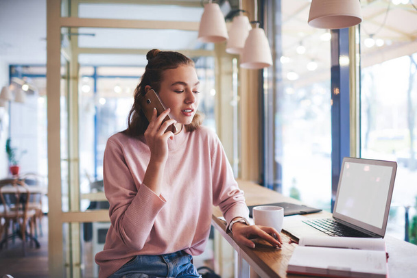 Young thoughtful female remote worker in casual clothes looking down sitting at table with cup of coffee and opened laptop while talking on phone - Photo, image