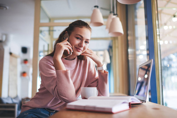 Happy female remote worker in casual outfit looking at camera sitting at table near window with opened laptop and cup of coffee while talking on phone during work - Foto, Imagem