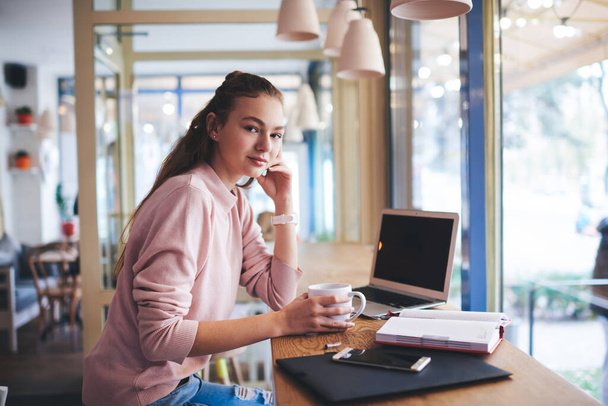 Side view of young calm female freelancer in casual outfit looking at camera sitting at table with laptop and cup of coffee having break - Fotó, kép