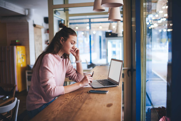 Side view of young female freelancer in casual clothes sitting at table with laptop and talking on smartphone while working on new project in cafe - Photo, image