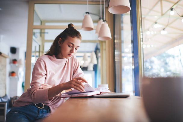 Focused female in casual outfit sitting at wooden table near window while reading interesting book and flipping page resting in light cafe - Foto, imagen