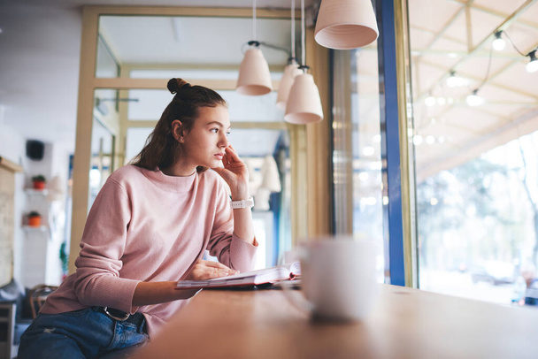 Young calm female in casual clothes looking away sitting at table with opened book and cup of coffee touching face and reading - Foto, immagini