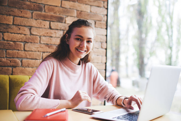 Positive female remote worker in casual clothes with toothy smile looking at camera sitting at table with opened laptop and doing work task - Foto, afbeelding