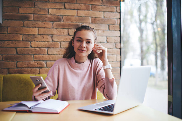 Young glad female remote worker in casual clothes looking at camera sitting at table with opened laptop touching face and surfing smartphone - Foto, immagini