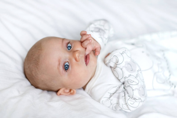 A little baby lying on the bed on a white sheet at home. Close up portrait of newborn baby looking at camera - Zdjęcie, obraz