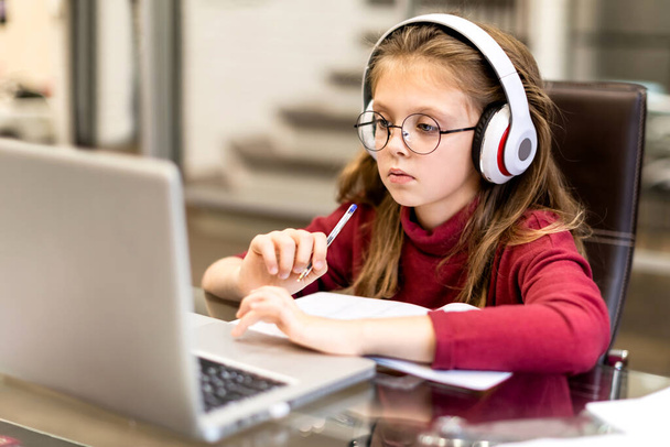 Cute little girl in wireless headphones seriously doing homework using laptop at home for online education and home studying. Kids distance learning. Social distance. Stay at home. - Fotó, kép