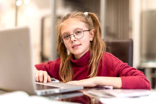 Evde online eğitim ve evde ders çalışmak için dizüstü bilgisayar kullanan mutlu küçük kız evde gülümseyerek kameraya bakıyor. Çocuklar uzaktan öğreniyor. Sosyal uzaklık. - Fotoğraf, Görsel