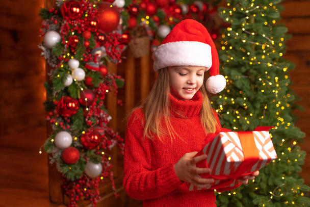 Happy child in a Santa red hat is holding Christmas gift with smile. Christmas concept. - Foto, afbeelding