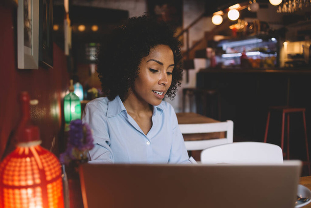 Young black focused female with curly hair in bluish shirt using laptop while sitting at wooden table in stylish cafe - Fotoğraf, Görsel