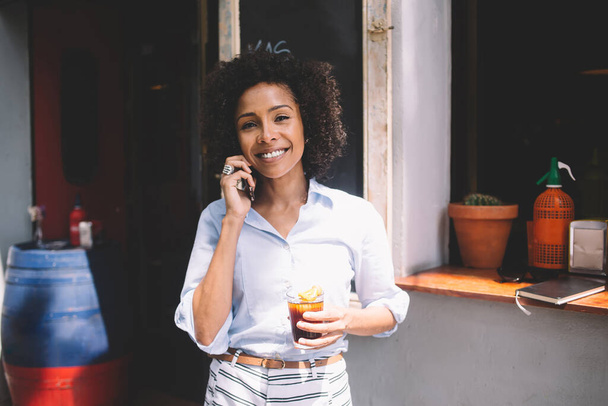 Young happy romantic African American female with curly hair talking on mobile phone and holding glass with drink while looking at camera - Фото, изображение