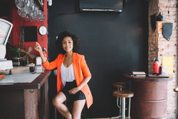 Confident young ethnic lady in stylish casual outfit sitting on chair near counter in bar with glass of fresh beverage and looking at camera - Zdjęcie, obraz