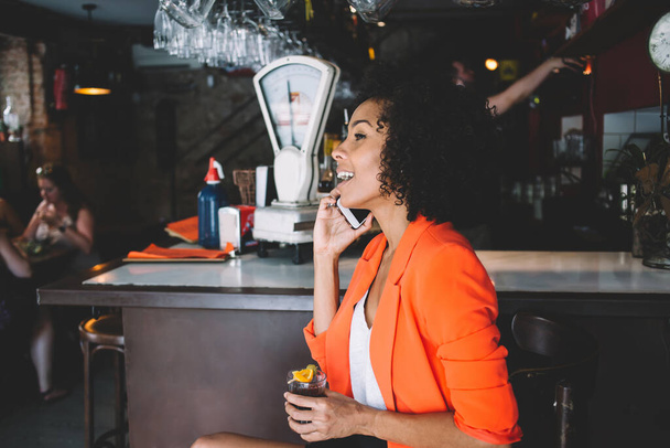 Side view of positive young ethnic lady speaking with friend on smartphone while sitting on chair and drinking cocktail in modern bar - Foto, Imagem